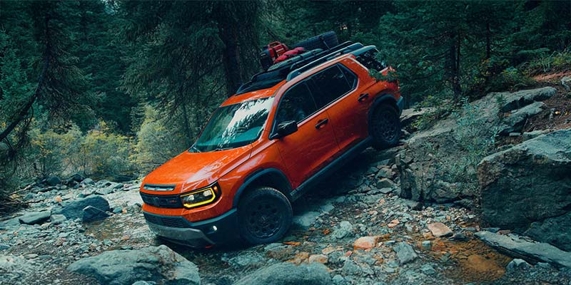 Orange SUV driving over rocky forest terrain.